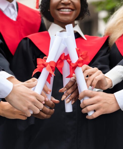 Cropped of multiracial group of students in graduation caps and robes posing at university campus, joining together their diplomas, celebrating graduation, closeup. Graduation concept