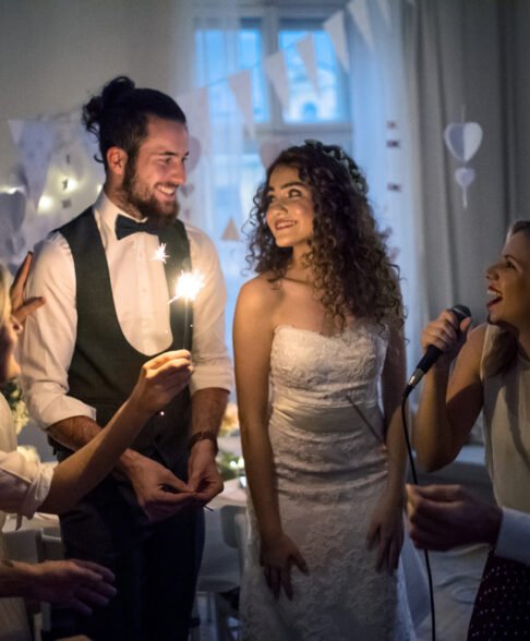 A young cheerful bride and groom with other guests dancing and singing on a wedding reception.