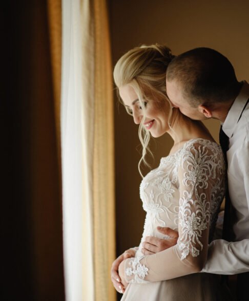 Bride and groom in a hotel room. The groom hugs and kisses the bride in the neck.