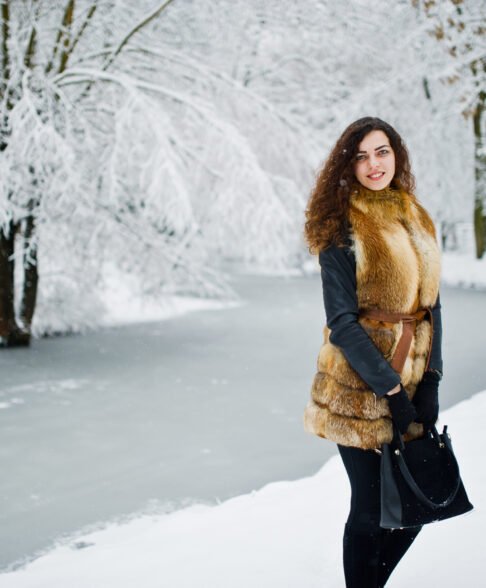 Elegance curly girl in fur coat at snowy forest park at winter.