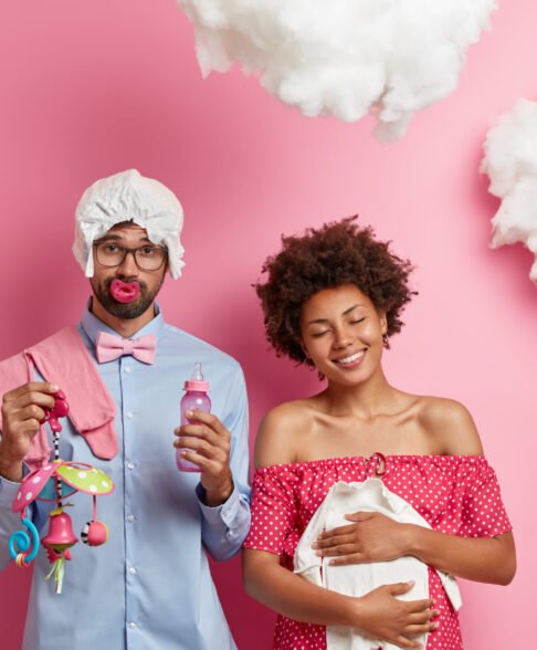 Happy dark skinned woman holds babys clothes over tummy smiles gently keeps eyes closed awaits for child. Sad future father with diaper on head keeps nipple in mouth holds feeding bottle mobile toy