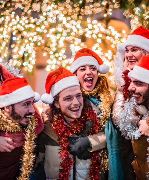 Happy group of friends wearing Santa Claus hat and celebrating Christmas night together - Young people having fun during winter holidays - Teenagers celebrating New Year's Eve in front of tree