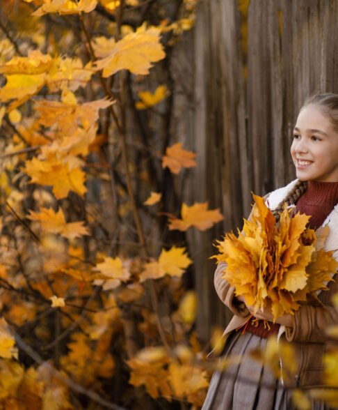 Smiling child girl standing near a wooden fence surrounded by yellow autumn leaves holding a large maple bouquet in her hands