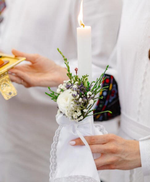 Woman wearing traditional embroidered shirt holding burning candle decorated with flowers during a religious ceremony