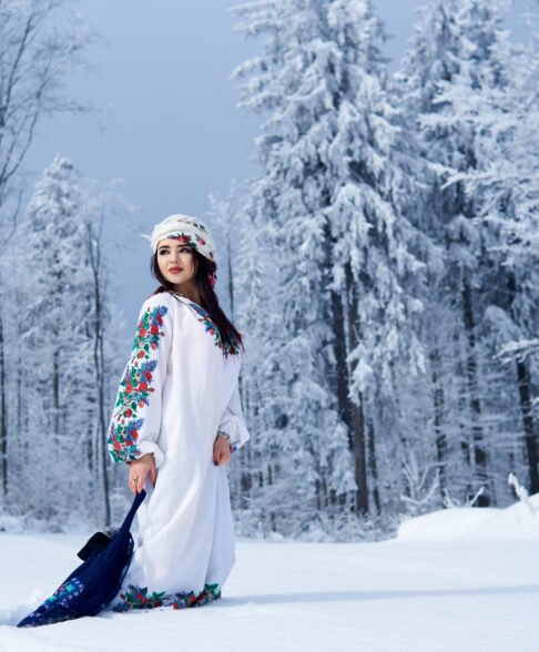 Young attractive fashionable dark-haired woman in white dress embroidered with floral design, colorful kerchief, with blue linen shoulder bag in deep snow outdoors on spruce forest winter background.